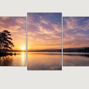Op de afbeelding: Een vijfdelige wandkunstset met een panoramisch landschap van een meer bij zonsondergang. De scène toont silhouetten van bomen op de linkeroever, een heldere oranje zon aan de horizon en een lucht vol zachte paarse en oranje wolken in het water.
