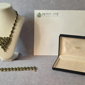 May include: A gold-tone necklace and bracelet set featuring oval dark green cabochon stones. The necklace is displayed on a bust, and the bracelet lies flat. An open black jewelry box and a white envelope with the text MASON KAY are included.