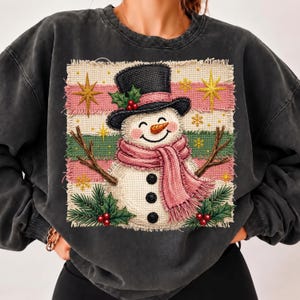 Puede incluir: Una sudadera de cuello redondo gris carbón con un gráfico cuadrado central de un muñeco de nieve con sombrero de copa negro y bufanda rosa. El muñeco tiene tres botones negros, nariz de zanahoria y está rodeado de estrellas doradas.