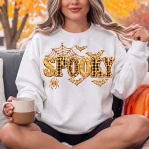 Peut inclure: Un sweat-shirt blanc à manches longues arborant le mot SPOOKY en grandes lettres décoratives avec des motifs dorés, à carreaux noirs et imprimé léopard. Le design inclut une toile d'araignée, une araignée et plusieurs chauves-souris dorées.