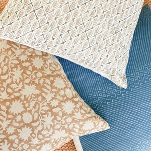 May include: Three decorative throw pillows with different patterns. The top pillow has a white background with a repeating pattern of small brown leaves. The middle pillow has a tan background with a repeating pattern of white flowers. The bottom pillow has a blue background with a repeating pattern of white geometric shapes.