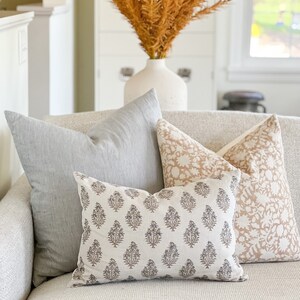 May include: Three decorative throw pillows on a light gray couch. The pillows are all rectangular and have different patterns. The pillow in the front is a light gray with a dark gray floral pattern. The pillow behind it is a light pink with a white floral pattern. The pillow in the back is a light gray with a subtle striped pattern.