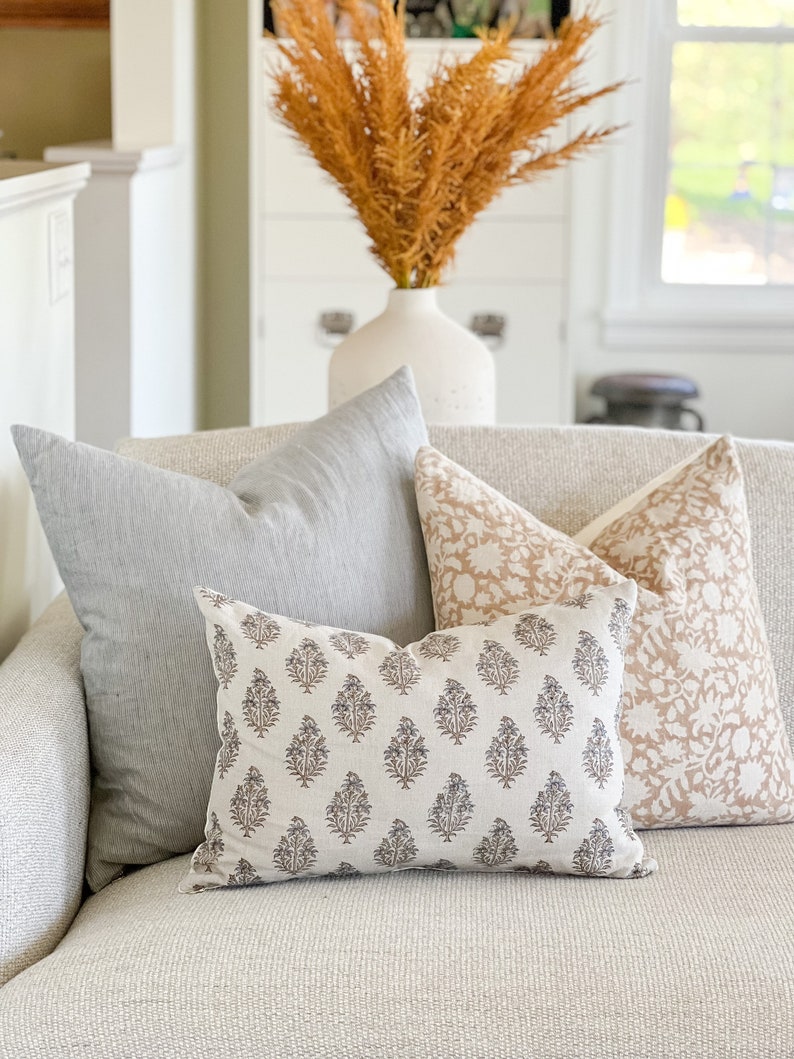 May include: Three decorative throw pillows on a light gray couch. The pillows are all rectangular and have different patterns. The pillow in the front is a light gray with a dark gray floral pattern. The pillow behind it is a light pink with a white floral pattern. The pillow in the back is a light gray with a subtle striped pattern.