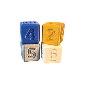 May include: Four stacked cubic blocks with embossed numbers. The top left block is blue with the number 4, the bottom left is silver with the number 5, the top right is gold with the number 2, and the bottom right is white with the number 6.