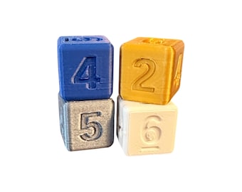 May include: Four stacked cubic blocks with embossed numbers. The top left block is blue with the number 4, the bottom left is silver with the number 5, the top right is gold with the number 2, and the bottom right is white with the number 6.