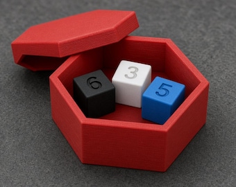 May include: A red hexagonal container with a matching lid, containing three small cubes. The black cube is marked with the number 6, the white cube with the number 3, and the blue cube with the number 5. The container has a textured, matte finish.