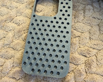 May include: A metallic blue-grey smartphone case featuring a textured surface with a uniform pattern of circular cutouts. The case has a large rectangular opening at the top for a camera module and a rigid, durable construction.