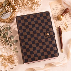 May include: A rectangular planner with a brown and black checkered pattern, featuring a snap button closure. The planner is accompanied by a dark wood fountain pen, a wax seal stamp, two gold paper clips, and decorative floral elements.