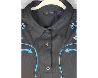 May include: A black button-down shirt featuring bright blue piping accents and embroidered cross designs on the chest. The garment includes a collar, a front placket with diamond-shaped metal buttons, and a Wrangler brand label at the neck.