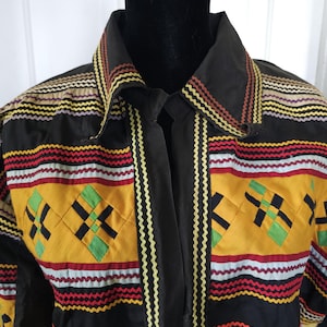 May include: A black button-down shirt featuring horizontal bands of yellow, red, and white fabric. The shirt is decorated with rows of multicolored rickrack trim and diamond-shaped patches in green, yellow, and black across the chest area.
