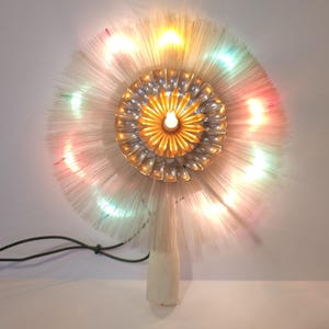 May include: A circular fibre optic tree topper featuring a central gold and silver metallic flower-like ornament. The design includes radiating white fibre optic strands illuminated by multi-coloured lights in shades of pink, blue, and yellow.