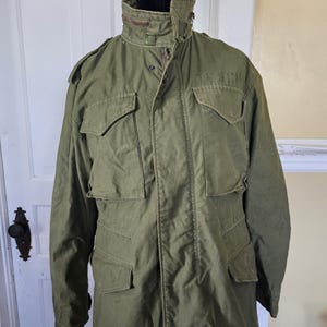 May include: An olive green field jacket featuring a high collar with a zipper detail, a front button placket, and four large flap pockets on the chest and waist. The jacket includes shoulder epaulets and the text U.S. ARMY printed on the chest.