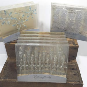 May include: Three Hallmark brand Christmas ornament sets in clear plastic packaging. One box contains blue beaded snowflake ornaments, one contains a clear snowflake garland, and the front box holds a set of twelve Glistening Icicles ornaments.