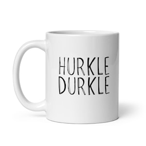May include: A white ceramic mug featuring the black text HURKLE DURKLE printed in a hand-drawn style font on the front. The mug has a smooth finish, a rounded handle on the left side, and a cylindrical body with a flat base for stability.