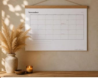 May include: A wall-mounted 2027 monthly calendar featuring a wooden hanging frame at the top. The white paper calendar displays the month of November with a grid layout, black text, and thin lines. It includes a section for notes at the bottom.