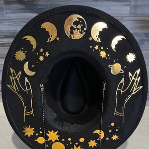 Pu&ograve; includere: Cappello fedora in feltro nero con una catena dorata e un design celeste. Il cappello presenta lune, stelle, pianeti e mani con accenti dorati.