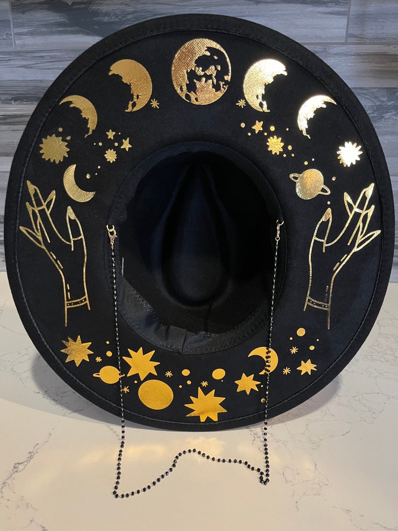 Pu&ograve; includere: Cappello fedora in feltro nero con una catena dorata e un design celeste. Il cappello presenta lune, stelle, pianeti e mani con accenti dorati.