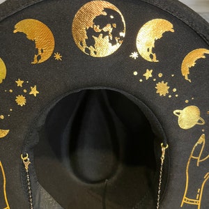 Pu&ograve; includere: Cappello fedora in feltro nero con disegni celesti dorati, tra cui lune, stelle e un pianeta. Il cappello ha una catena dorata attaccata al bordo.