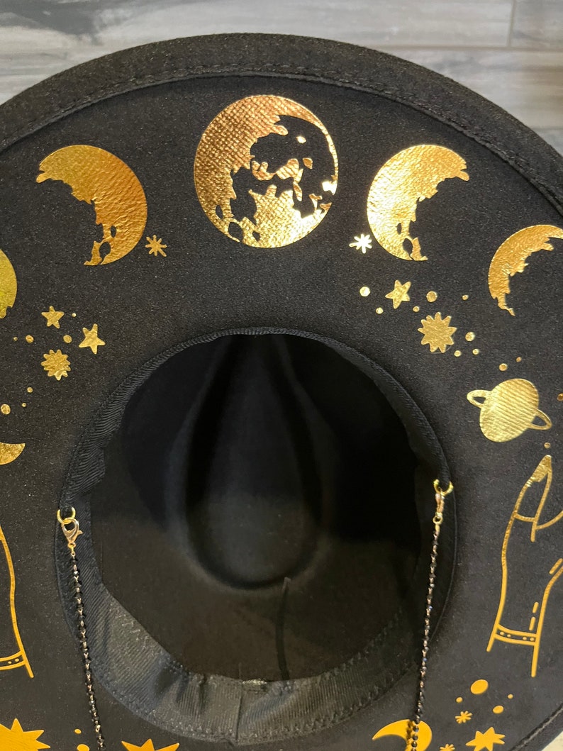 Pu&ograve; includere: Cappello fedora in feltro nero con disegni celesti dorati, tra cui lune, stelle e un pianeta. Il cappello ha una catena dorata attaccata al bordo.