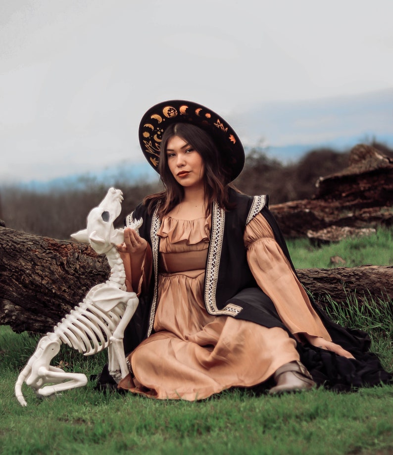 Pu&ograve; includere: Una donna con un mantello nero e un vestito marrone con un cappello nero con disegni celesti dorati &egrave; seduta su un tronco con un cane scheletro bianco al suo fianco.
