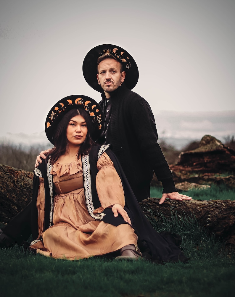 Pu&ograve; includere: Una coppia indossa cappelli neri con disegni dorati di luna e stelle. La donna indossa un vestito marrone con un mantello nero e l'uomo indossa una giacca nera. Sono in piedi davanti a un tronco d'albero.