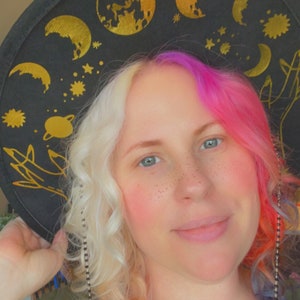 Pu&ograve; includere: Una donna con capelli biondi e rosa indossa un cappello nero con disegni celesti dorati. Il cappello ha una catena.
