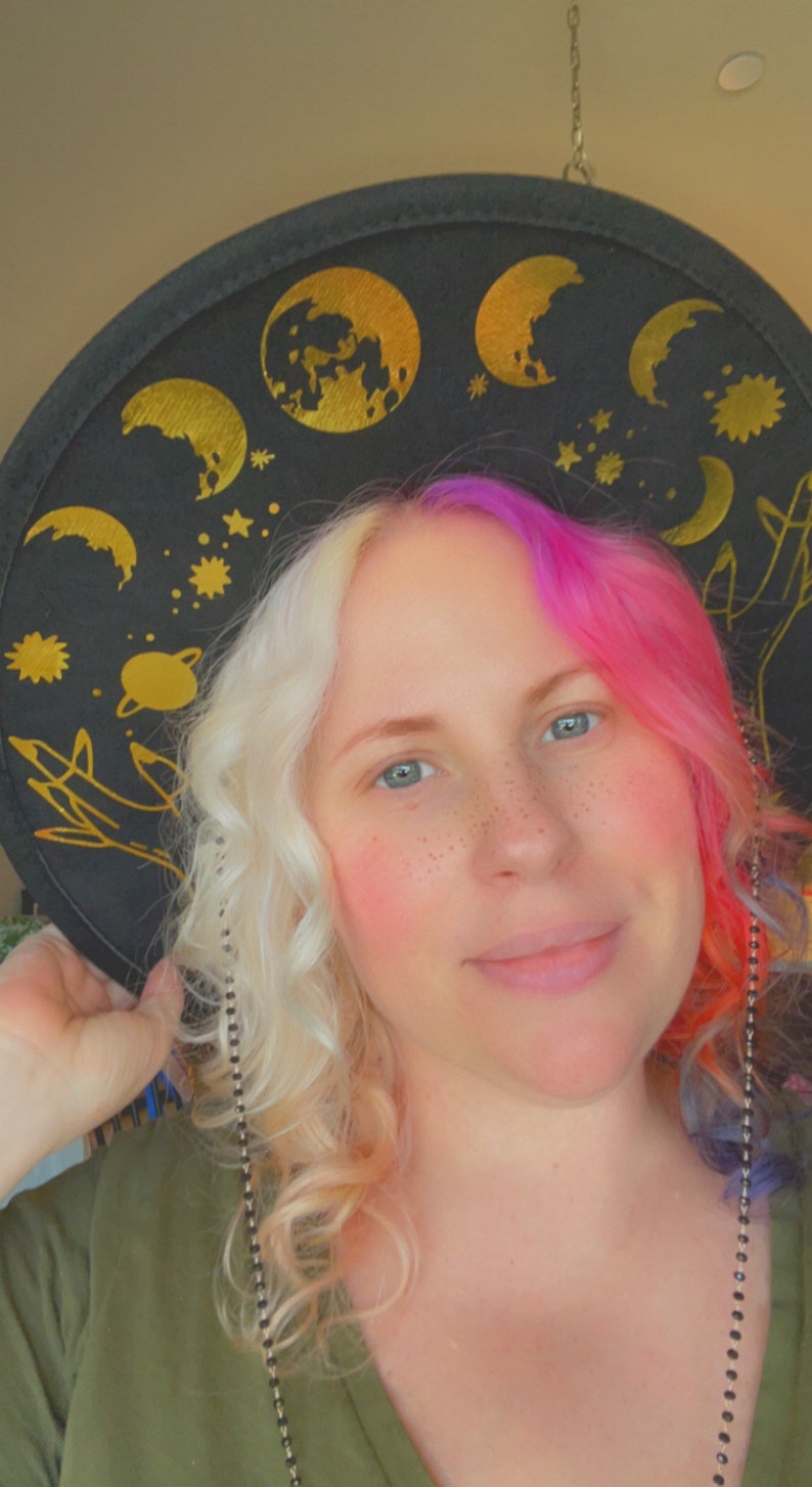 Pu&ograve; includere: Una donna con capelli biondi e rosa indossa un cappello nero con disegni celesti dorati. Il cappello ha una catena.