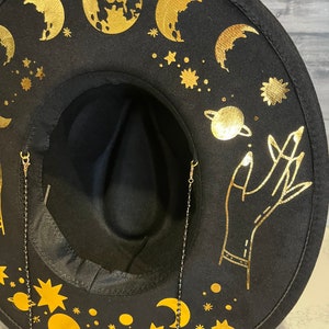 Pu&ograve; includere: Cappello fedora in feltro nero con disegni celesti dorati, tra cui lune, stelle e una mano che raggiunge un pianeta. Il cappello ha due catene nere attaccate al bordo.
