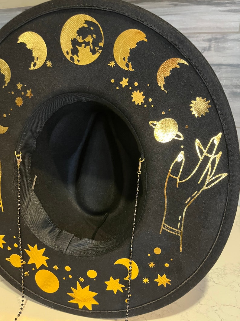 Pu&ograve; includere: Cappello fedora in feltro nero con disegni celesti dorati, tra cui lune, stelle e una mano che raggiunge un pianeta. Il cappello ha due catene nere attaccate al bordo.
