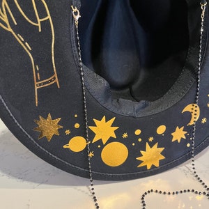Pu&ograve; includere: Un cappello fedora in feltro nero con un design celeste dorato, che include una luna crescente, stelle e pianeti. Il cappello ha una catena di perline nere attaccata all'interno.