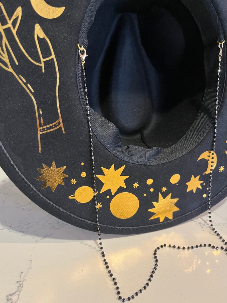 Pu&ograve; includere: Un cappello fedora in feltro nero con un design celeste dorato, che include una luna crescente, stelle e pianeti. Il cappello ha una catena di perline nere attaccata all'interno.