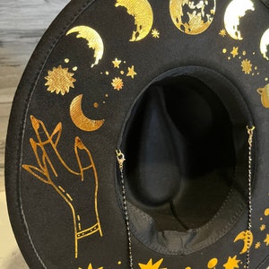 Pu&ograve; includere: Cappello fedora in feltro nero con un design celeste dorato. Il cappello presenta lune, stelle, pianeti e una mano alzata. Il cappello ha una catena nera attaccata al bordo.
