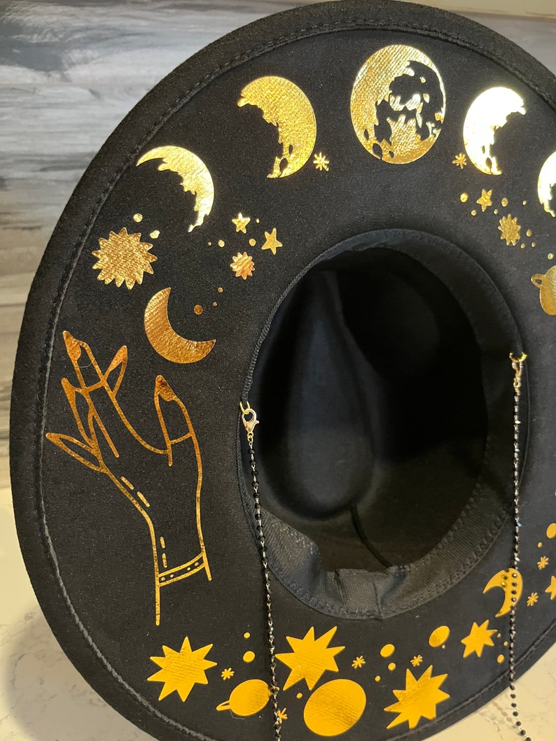 Pu&ograve; includere: Cappello fedora in feltro nero con un design celeste dorato. Il cappello presenta lune, stelle, pianeti e una mano alzata. Il cappello ha una catena nera attaccata al bordo.