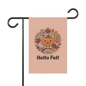 Puede incluir: Una bandera de jardín rectangular en color melocotón suave con una ilustración central de una calabaza naranja sonriente con un lazo a cuadros, rodeada por una corona de hojas de otoño, bellotas y piñas, con el texto Hello Fall debajo.