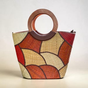 Pode incluir: Uma bolsa com corpo de palha trançada e um padrão de patchwork de segmentos curvos em vermelho, laranja e bege. A bolsa possui uma alça circular de madeira fixada a um acabamento de aspecto de couro marrom escuro na parte superior.