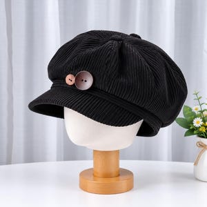 May include: A black corduroy newsboy cap featuring a textured ribbed fabric and a short, stiff brim. Two round, brown buttons of different sizes are attached to the front band of the cap, providing a contrasting detail against the dark material.
