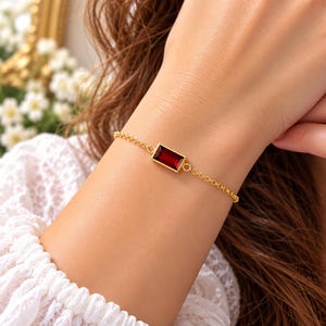 May include: A gold chain bracelet featuring a single rectangular, deep red garnet gemstone set in a gold bezel. The bracelet consists of delicate, circular gold links that connect to the central stone setting, creating a simple and elegant design.