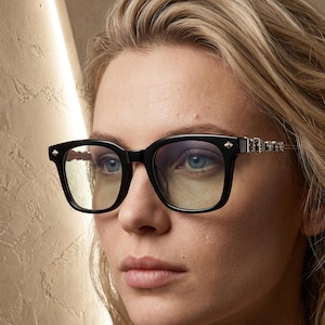 May include: Black-framed spectacles with a thick, rectangular shape and clear lenses. The temples feature intricate silver-toned metal detailing with a small, pointed stud accent at the outer corners of the frame front, providing a distinct look.
