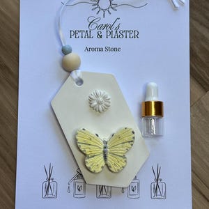 May include: A white tag-shaped aroma stone featuring a yellow butterfly and a small white daisy. It hangs from a white ribbon with a blue and a wooden bead. A small glass dropper bottle with a gold collar is included on the branded display card.