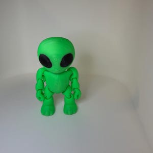 May include: A bright green articulated alien figurine with a large, smooth, rounded head and two prominent, oval-shaped black eyes. The figure features a slender torso, jointed arms, and legs, standing upright with a distinct, textured surface finish.