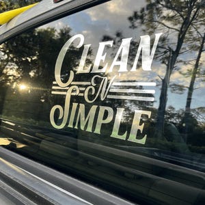 May include: A silver-colored vinyl decal applied to a vehicle window featuring the text "CLEAN N' SIMPLE" in a stylized font. The word "CLEAN" is on the top line, "N'" is in the middle with horizontal lines, and "SIMPLE" is on the bottom line.