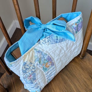 May include: A quilted fabric pouch featuring a patchwork design of white, blue, and yellow floral and striped patterns. The pouch has a bright light blue interior lining and a matching light blue fabric bow tied at the top center opening.