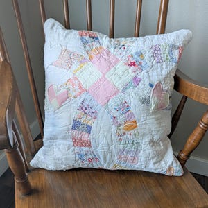 May include: A square throw pillow featuring a white background with a central patchwork design of interlocking rings in various pastel floral and geometric patterns. Two small heart-shaped fabric appliqués are stitched onto the front surface.