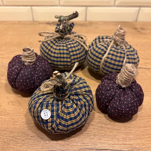 Op de afbeelding: Een verzameling van vijf stoffen pompoenen in blauw en geel ruitpatroon of donkerpaars met een cirkelvormig patroon. Elke pompoen heeft een steel van omwikkeld touw, sommige met kleine takjes, en één blauwe ruitpompoen heeft een witte knoop.