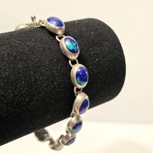 Peut inclure: Un bracelet à maillons en argent composé d'une série de pierres cabochons ovales bleu profond avec des inclusions bleu sarcelle. Chaque pierre est sertie dans une monture en argent lisse, reliée par des anneaux et un fermoir métallique.