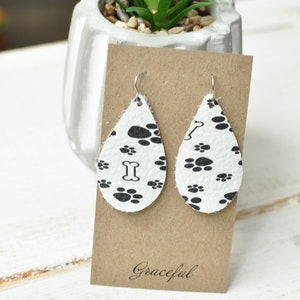May include: A pair of white leather teardrop earrings with a black paw print pattern. The earrings have silver hooks and a bone charm on one earring. The earrings are displayed on a brown card with the word "Graceful" written on it.