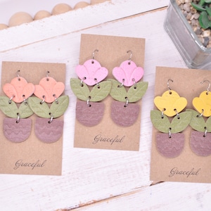 May include: Three pairs of dangle earrings with a flower and pot design. The earrings are made of faux leather and come in pink, peach, and yellow. Each pair is displayed on a brown card with the word "Graceful" printed on it.