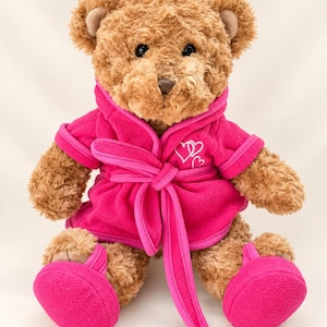 May include: A brown plush teddy bear wearing a bright fuchsia fleece bathrobe with a tie belt and matching fuchsia slippers. The bathrobe features two embroidered white heart outlines on the left chest area, and the bear has dark eyes.