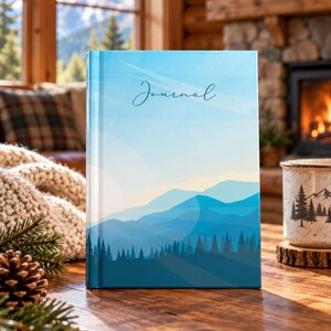 May include: A hardcover journal featuring a graphic illustration of layered blue mountains and a dark forest under a light blue sky. The word Journal is printed in a thin, dark script font centered near the top of the front cover.