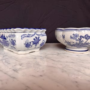 Puede incluir: Una jardinera rectangular de cerámica y un cuenco redondo de cerámica, ambos con base blanca y motivos florales y paisajísticos azules. La pieza rectangular tiene un borde festoneado, mientras que el cuenco redondo se asienta sobre una base.